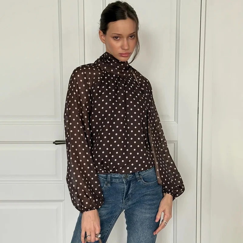 Woman wearing a brown polka dot blouse and blue jeans against a white paneled wall.