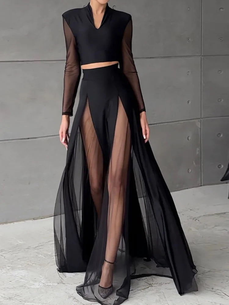Person wearing a black outfit with sheer elements against a gray wall.