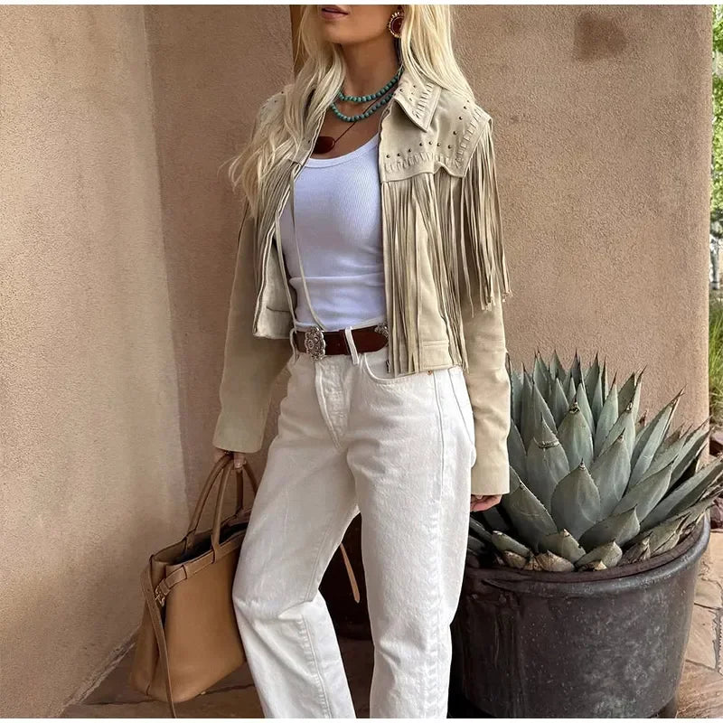 Person wearing a beige fringe jacket, white pants, and holding a beige handbag against a beige wall with a potted plant.