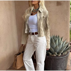 Person wearing a beige fringe jacket, white pants, and holding a beige handbag against a beige wall with a potted plant.