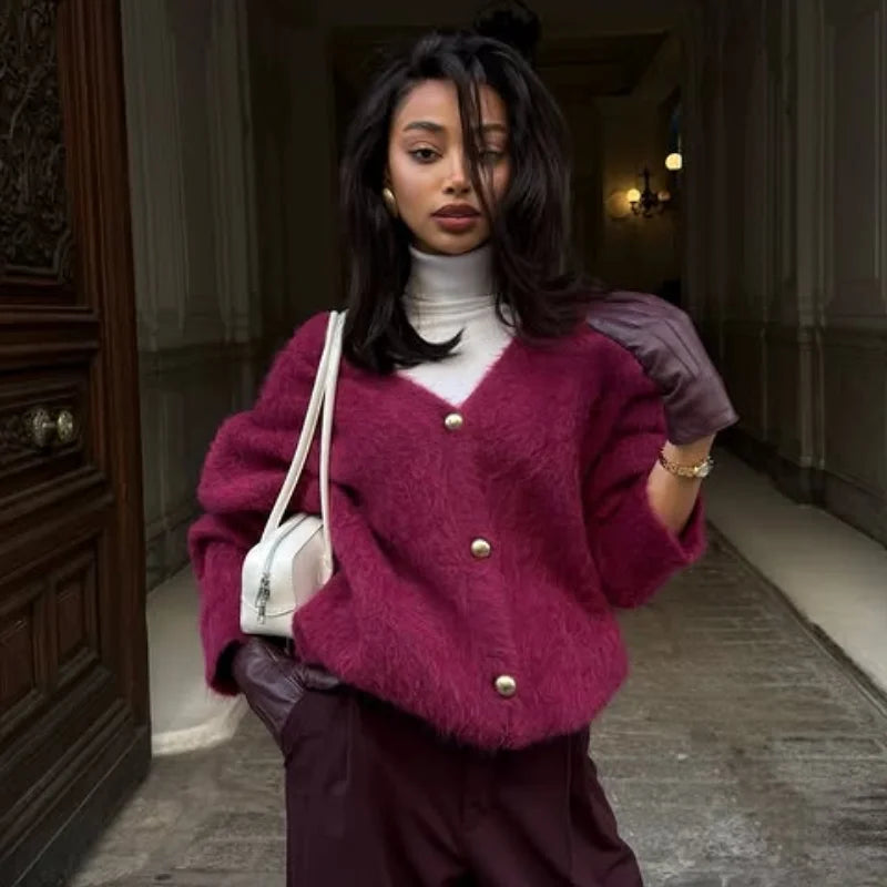 Woman wearing a pink cardigan with gold buttons, holding a white bag, standing in an indoor setting.