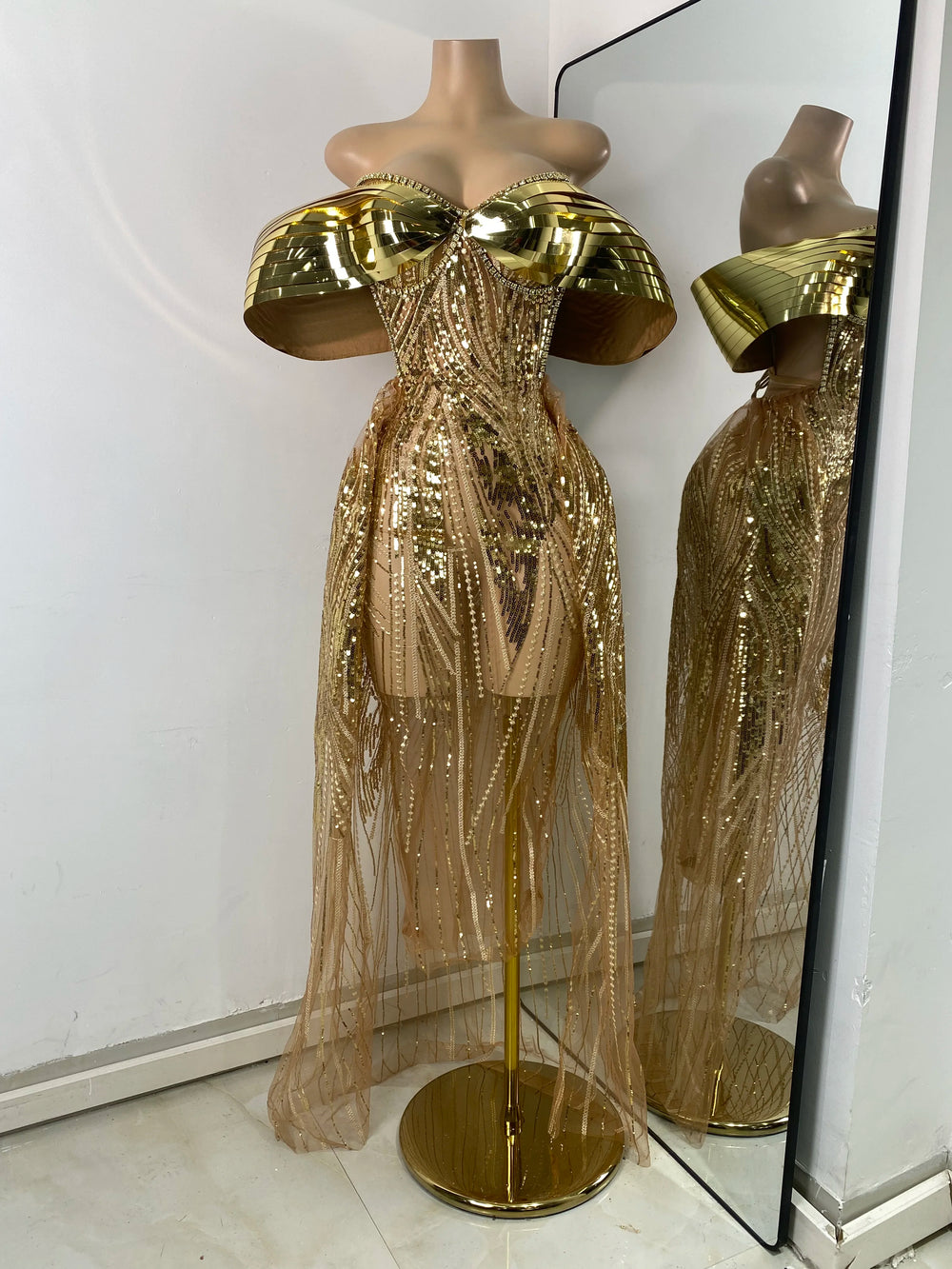 Gold sequin dress on a mannequin with a mirror reflection against a white wall.