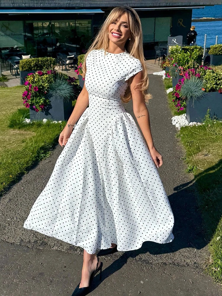 Woman in a white polka dot dress standing outdoors with flowers and water in the background