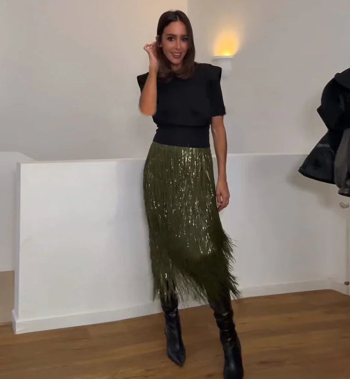 Woman wearing a black top and green sequin skirt in a room with white walls and wooden floor.