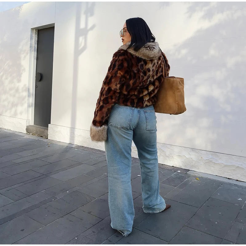 Person wearing a leopard print coat and blue jeans standing against a white wall.