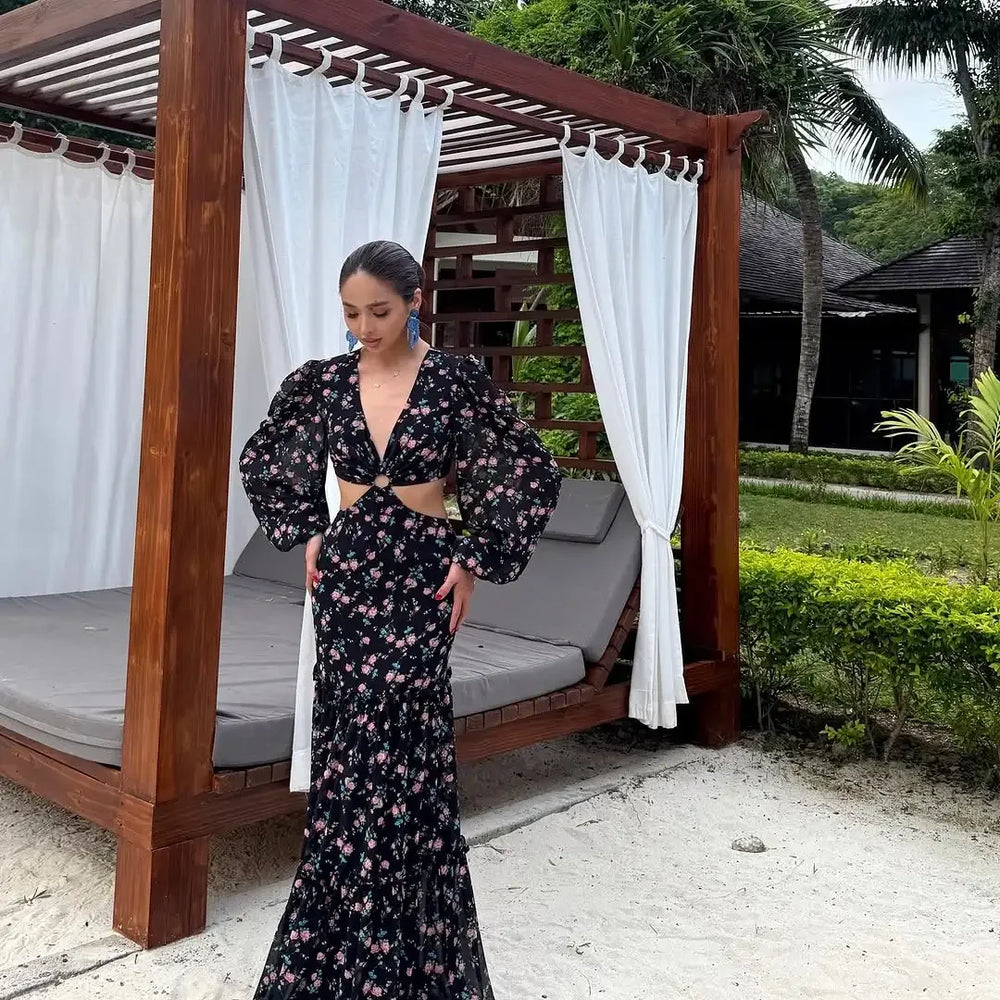 Woman in a floral dress standing under a wooden canopy with white curtains in a garden setting.