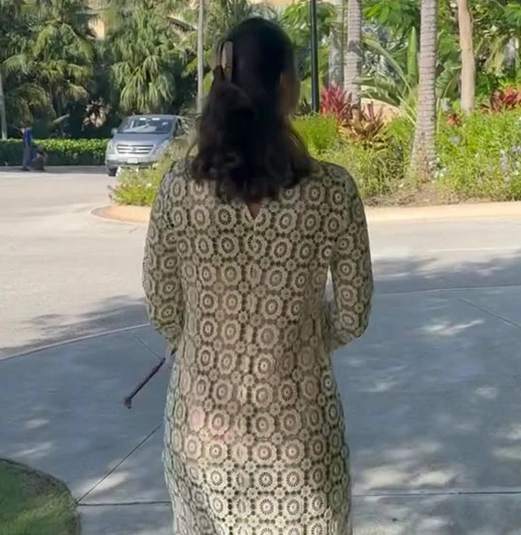 Woman in a patterned dress standing on a sidewalk with palm trees and a car in the background.