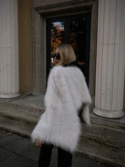 Person wearing a white fur coat standing in front of classical columns.