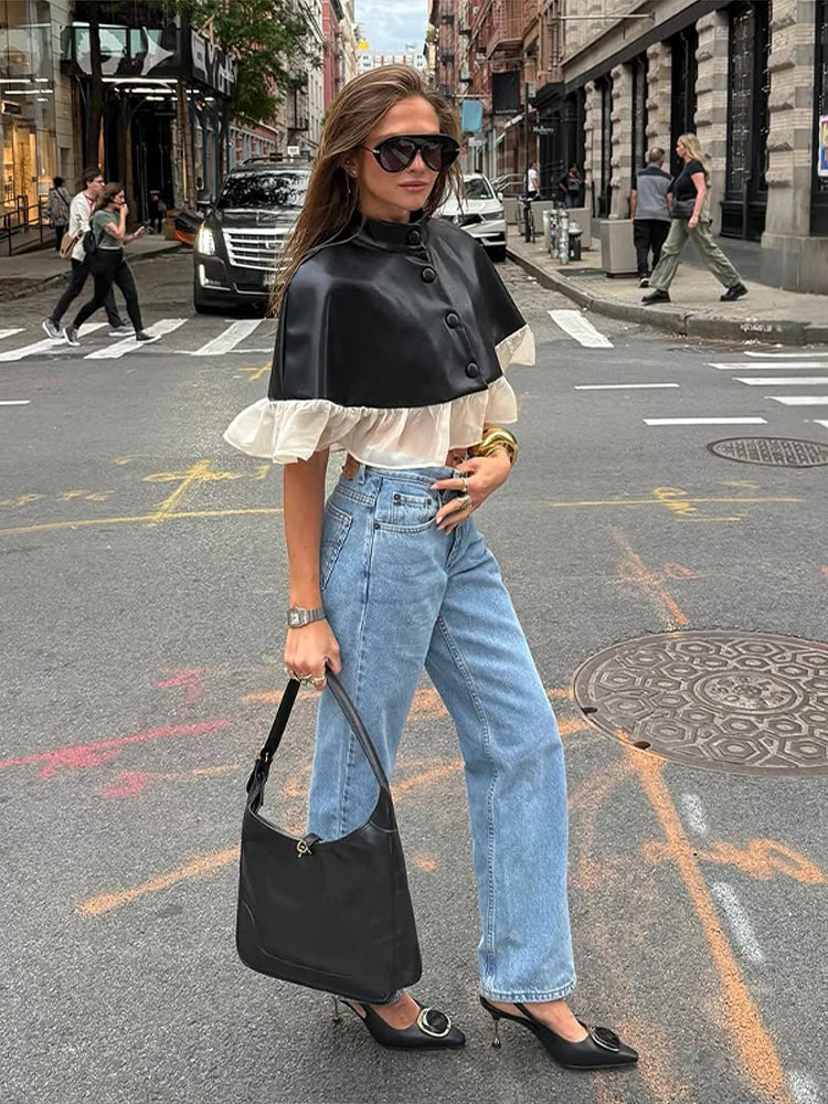 Woman in black leather jacket, white blouse, and blue jeans standing on a city street.