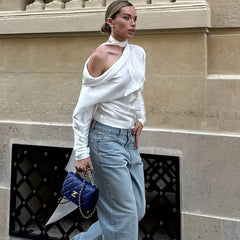 A woman in a Y2K inspired streetwear polyester slim fit top in white and light blue jeans holding a blue handbag against a beige wall.
