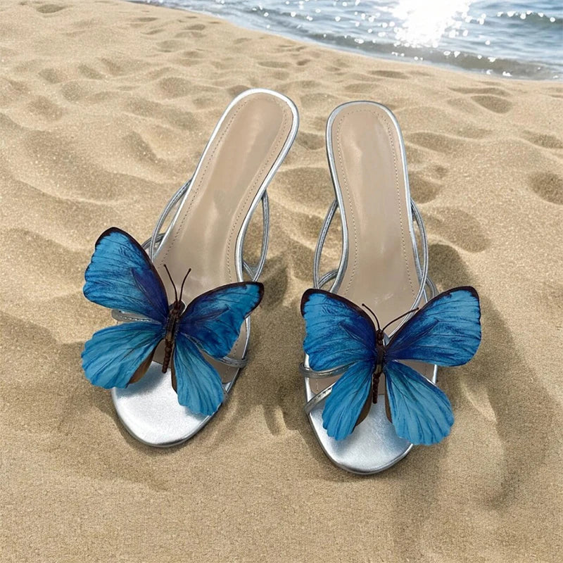 Silver high-heeled sandals with blue butterfly decorations on a sandy beach.