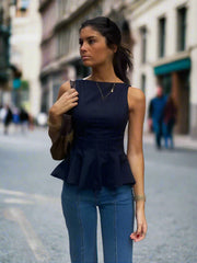 Woman wearing a back-zip peplum tank shown from the front with clean round neck and blue jeans standing  in a street.