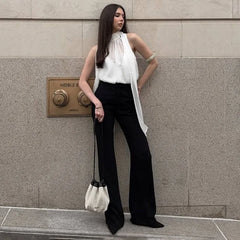 Woman in a backless, elegant clubwear top in white with bow detail for nightlife and black pants standing against a grey wall.