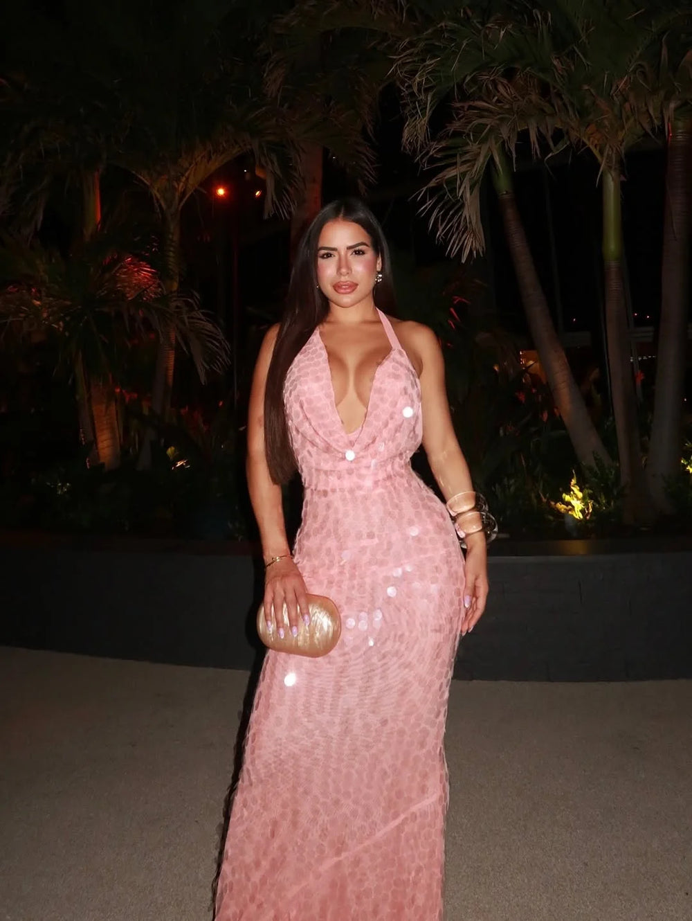 Woman in a backless, floor-length dress with lace-up detail and sparkle, standing outdoors at night with palm trees in the background