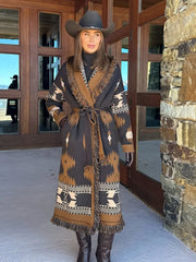 Full-length shot of a woman wearing a brown printed belted knit cardigan with tassel trim, styled with tall boots outside stone walls.