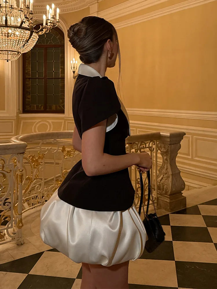 Side look of a black-and-ivory two-piece set with puff sleeves and a mini skirt, photographed in a grand interior with ornate railings.
