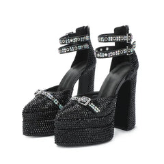 Front angle showing black open-toe platform sandals with a sparkling pearl bow detail and a buckle strap wrapping the ankle.