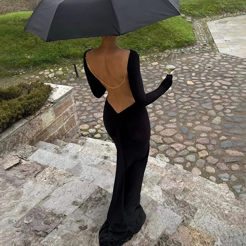 Back view of a black long sleeve mermaid maxi dress with an open back and fine chain detail, styled with an umbrella outdoors.