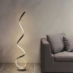 A modern black floating LED floor lamp with an illuminated golden spiral design, placed in a room with a grey wall and a sofa next to it.