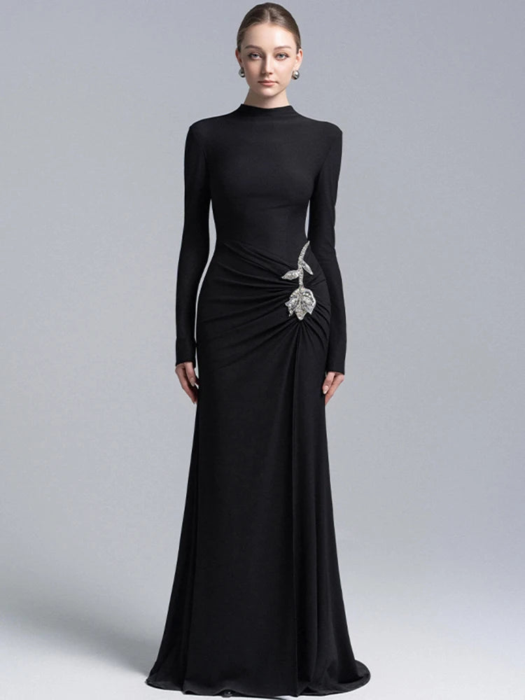 Model wearing a black high neck long sleeve maxi gown with draped waist and crystal floral detail, photographed on a clean studio background
