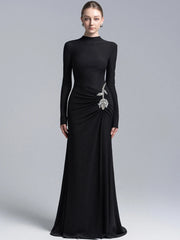 Model wearing a black high neck long sleeve maxi gown with draped waist and crystal floral detail, photographed on a clean studio background