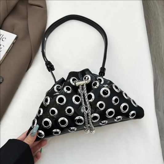 Black drawstring underarm bag on a white surface, covered in metal eyelets with chain accents beside a tailored blazer.