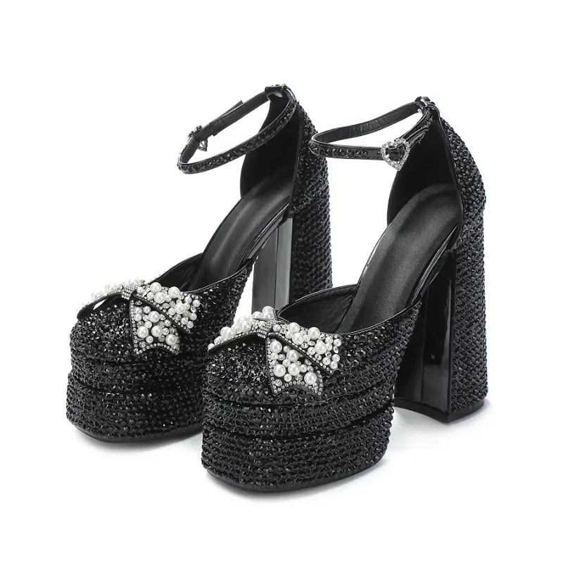 Pair of black platform block-heel sandals on a white background with pearl bow embellishments and ankle straps for a dressy finish.