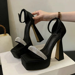 Black high-heeled shoes with rhinestone straps held by a hand on a white surface.
