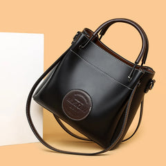 A black, soft PU leather crossbody handbag with three handles and a round logo on the front, displayed against a beige background.