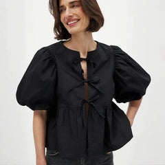 Model wearing a black tie-front peplum blouse with puff sleeves, smiling in a clean studio setting with soft, minimal styling.
