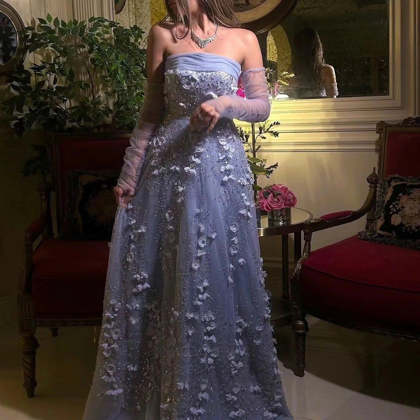 Woman in a blue evening gown standing in an elegant room with red chairs and floral decorations.