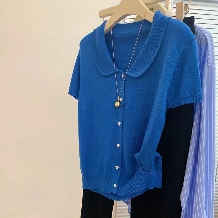 Blue cardigan with a gold necklace on a hanger against a beige wall.