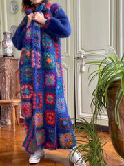 A person wearing a colourful, plaid, hand-crocheted long jumper with a geometric pattern, in rose red and royal blue, standing in a room with a potted plant and a wooden chair.