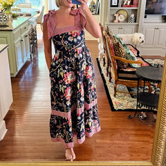 A woman taking a mirror selfie wearing a Boho floral swing dress with wide bow-tied shoulder straps in a home setting.