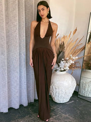 Full-length view of a brown deep V maxi dress with a fitted bodice and pleated skirt, styled in a softly decorated interior.