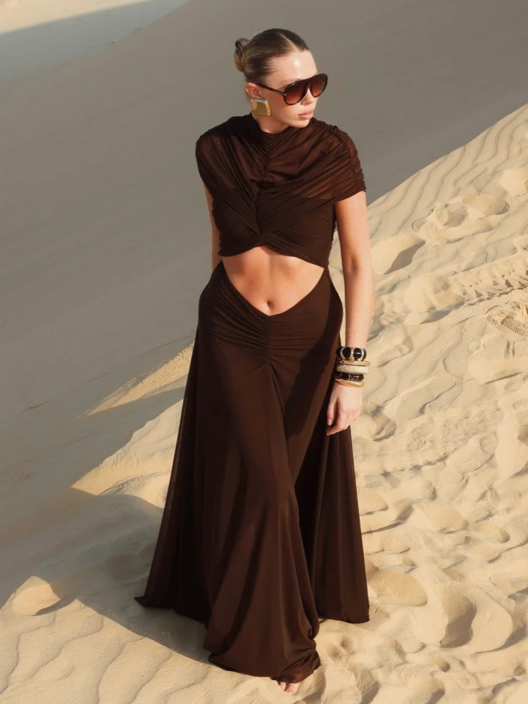 Model in a brown ruched maxi dress with a waist cutout, standing on sand dunes with sunglasses and stacked bracelets.