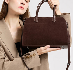 Model holding a brown suede leather shell bag at chest level, showing the structured front panel and smooth top handles.