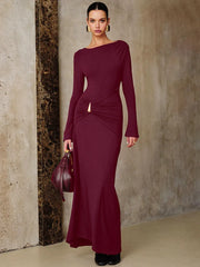 A Woman wearing a burgundy ruched long-sleeve maxi dress styled for an autumn party against a textured wall.