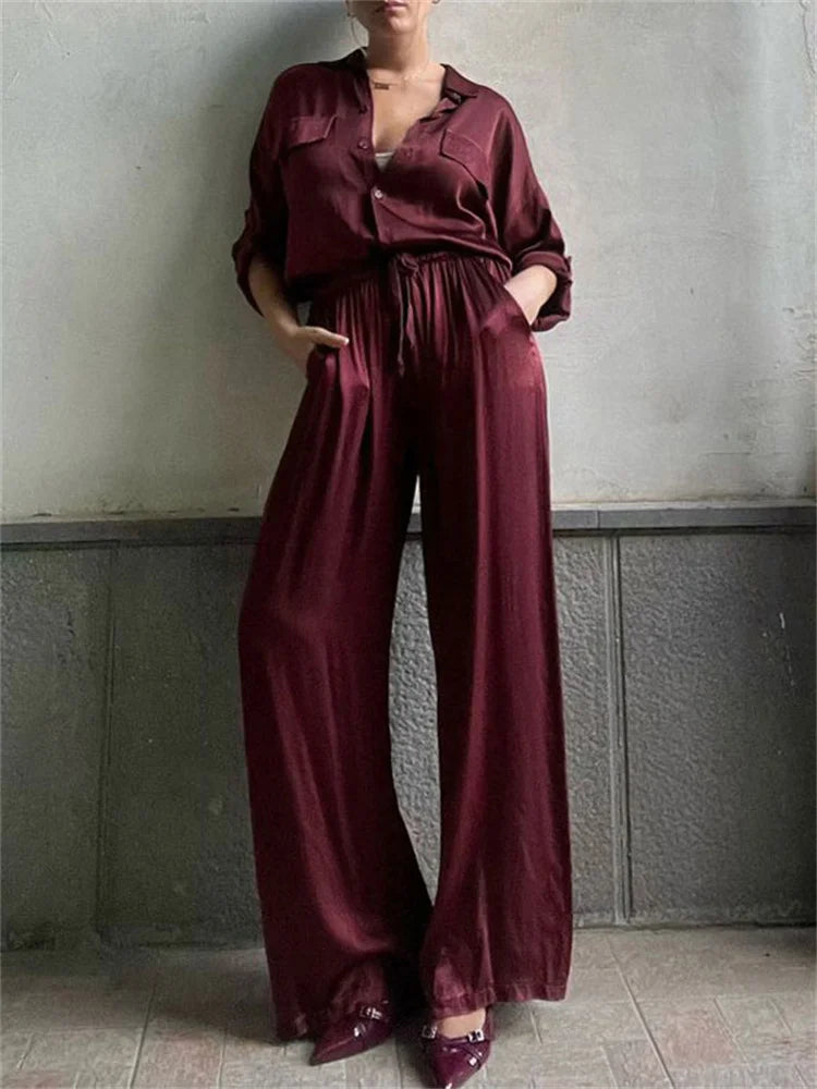 Person wearing a Burgundy satin jumpsuit set with wide-leg pants and button-up shirt against a plain wall.