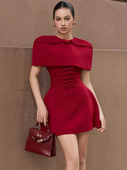 Model wearing a deep red cape sleeve corset mini dress, holding a structured top-handle bag against a textured wall with a polished stance.