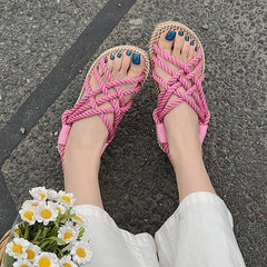 Pink braided sandals worn on a person's feet with a white floral clutch on a gray pavement background.