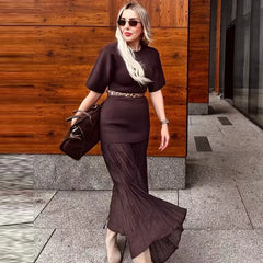 Woman in a dark brown dress holding a handbag against a wooden wall.