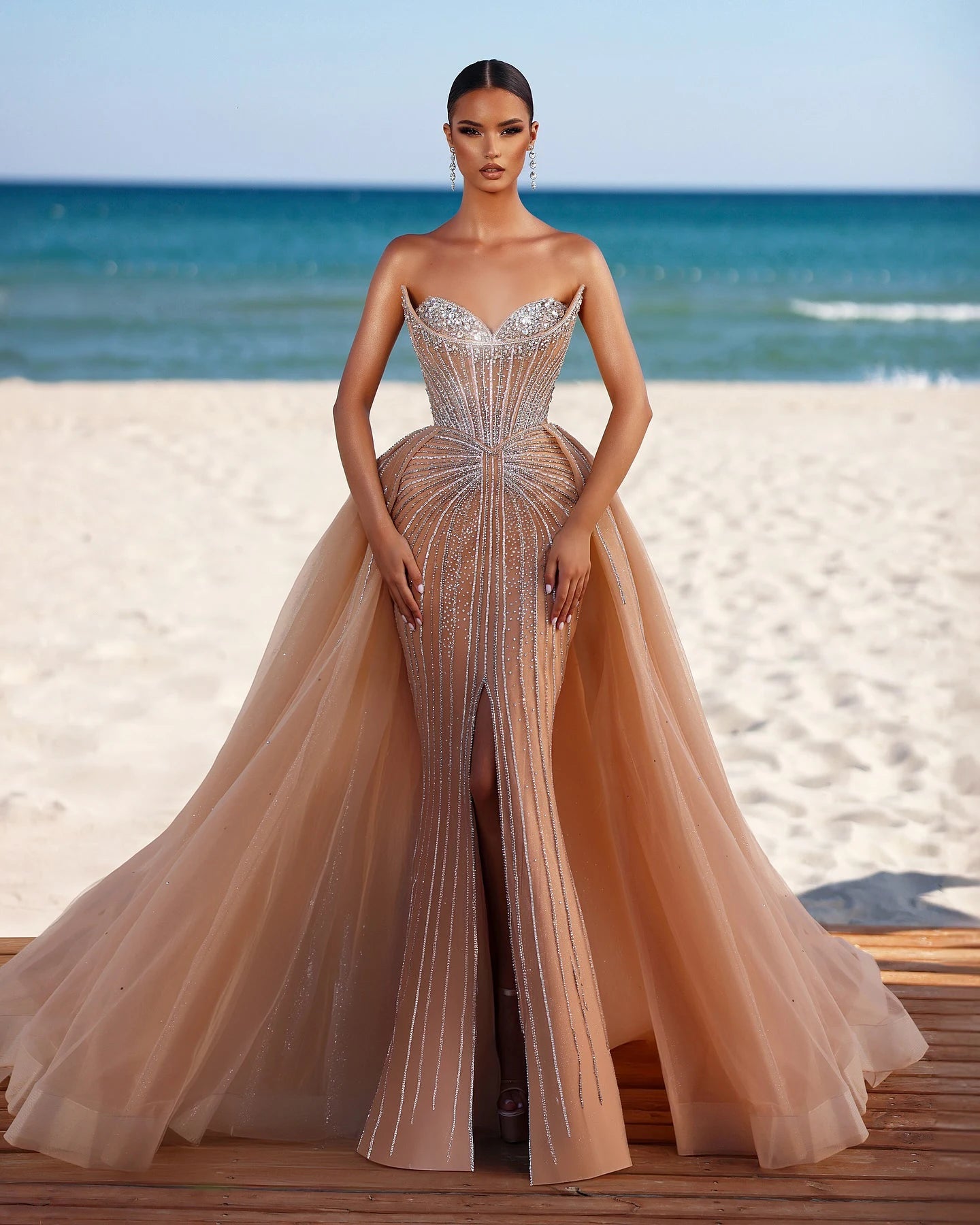 Woman in a glamorous champagne sweetheart mermaid gown with detachable tulle overskirt for weddings on a beach with ocean in the background