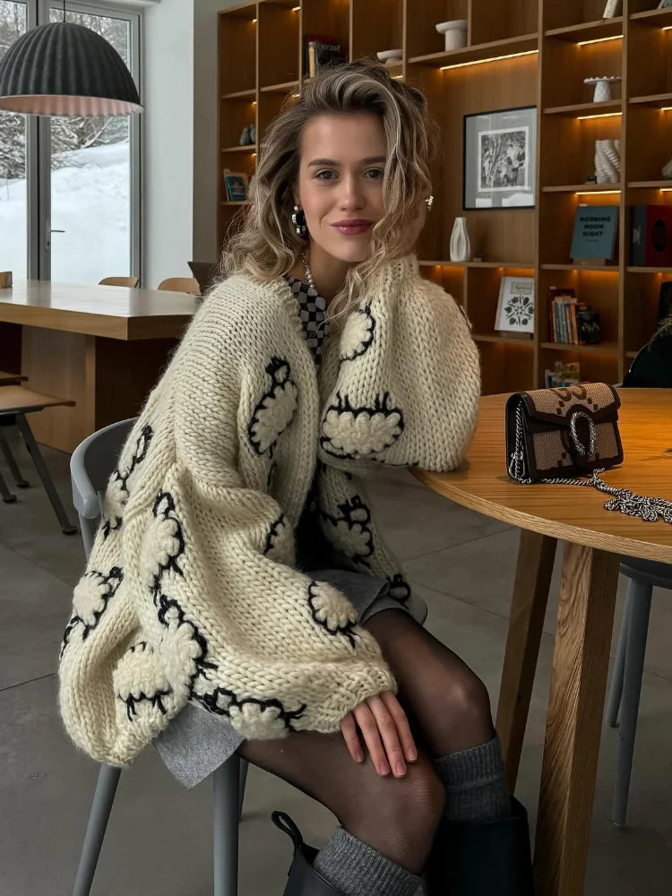 Woman wearing a cream-colored cardigan with black patterns sitting in a cozy room.