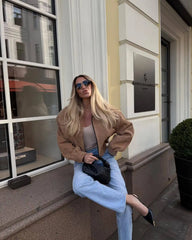 Woman sitting on a building ledge wearing sunglasses and a  cropped streetwear autumn coat in  khaki with a classic turn-down collar