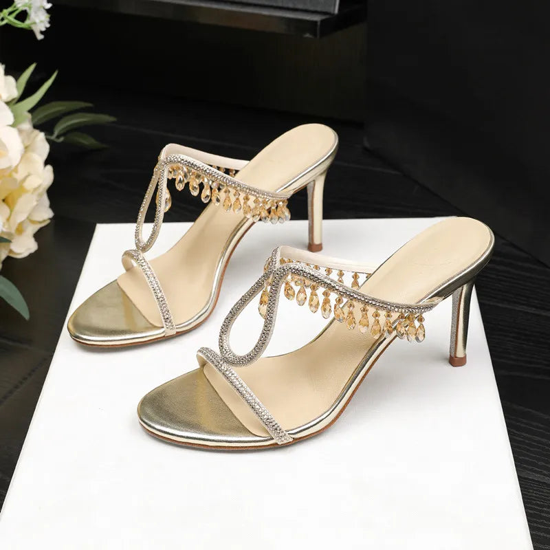 Crossed-leg styling shot featuring crystal-embellished open-toe heels, focusing on the strap sparkle and refined evening finish.