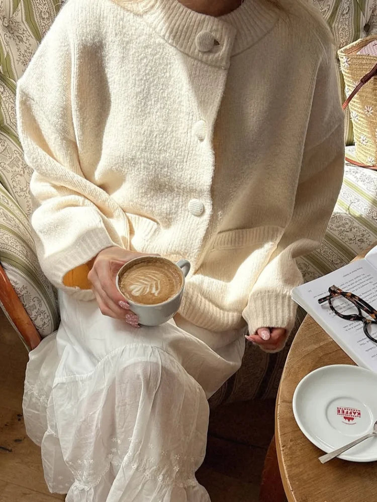 Person wearing a white cardigan holding a cup of coffee in a cozy setting.