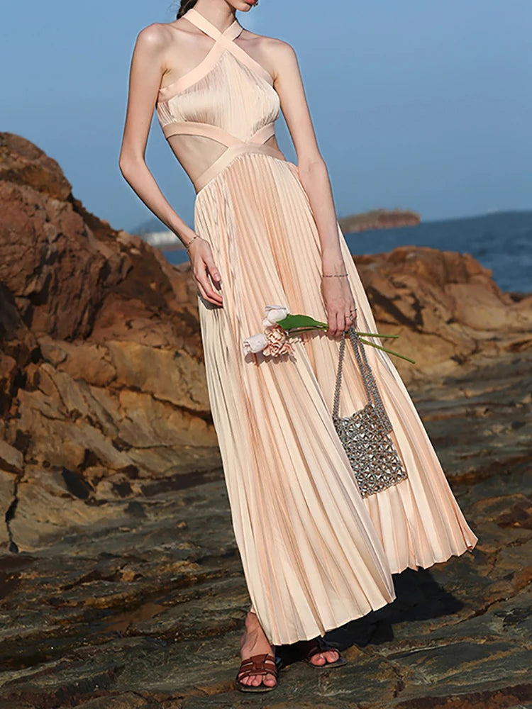 A woman in an elegant summer gown with waist cutouts and soft pleats is standing on a rocky beach with a clear blue sky.