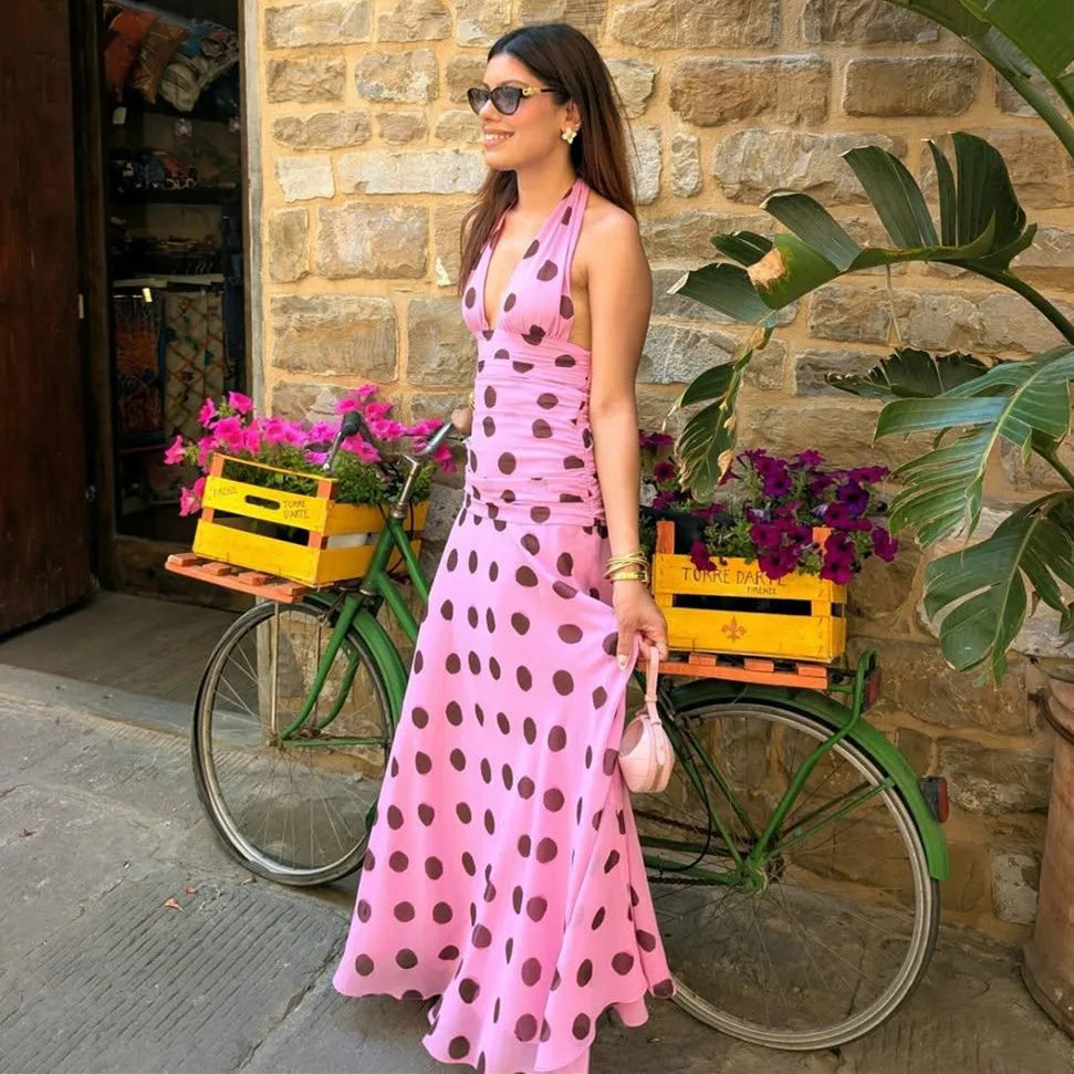 Woman in a pink polka dot dress standing next to a green bicycle with flower crates, against a stone wall.
