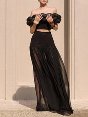 Woman wearing a black off-shoulder top and long skirt against a beige wall.